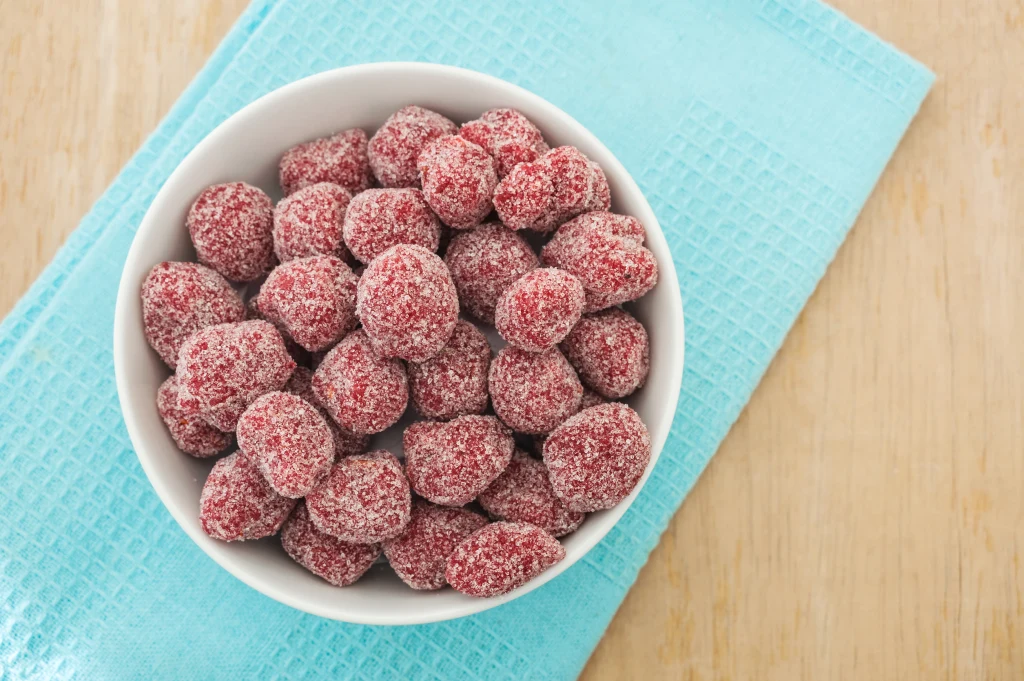 W krajach azjatyckich popularne są cukierki tamaryndowe przygotowywane z owoców i odrobiny przypraw Biała miska wypełniona czerwonymi żelkami obtoczonymi w cukrze, ustawiona na turkusowej ściereczce na jasnym drewnianym blacie.