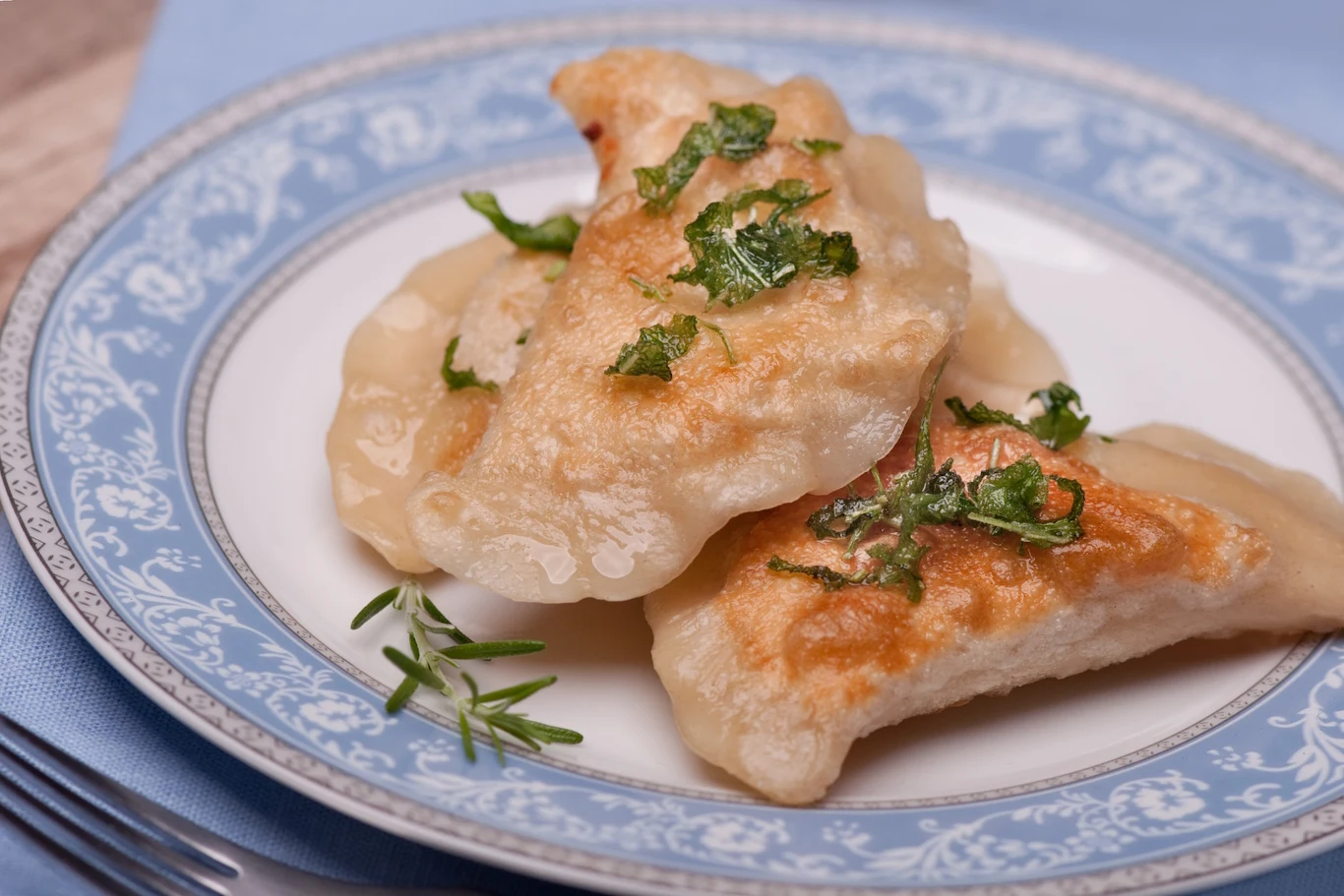 Farsz z twarogiem, serkiem topionym i ubitymi ziemniakami jest idealnie gładki i kremowy Trzy rumiane pierogi posypane ziołami leżące na eleganckim talerzu z dekoracyjnym wzorem, położonym na niebieskiej serwetce.