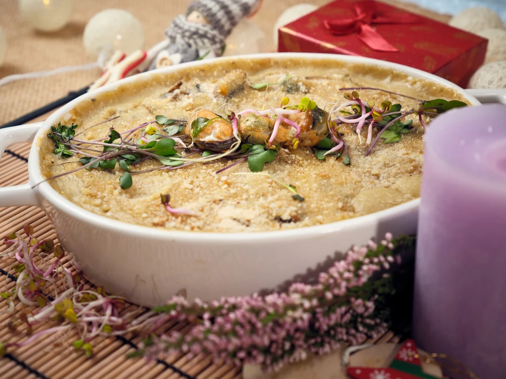 Zapiekanka w białym naczyniu ozdobiona świeżymi ziołami i kiełkami, ustawiona na dekoracyjnym stole, w tle fioletowa świeca, kwiat wrzosu oraz świąteczne dodatki i czerwone pudełko z kokardą.