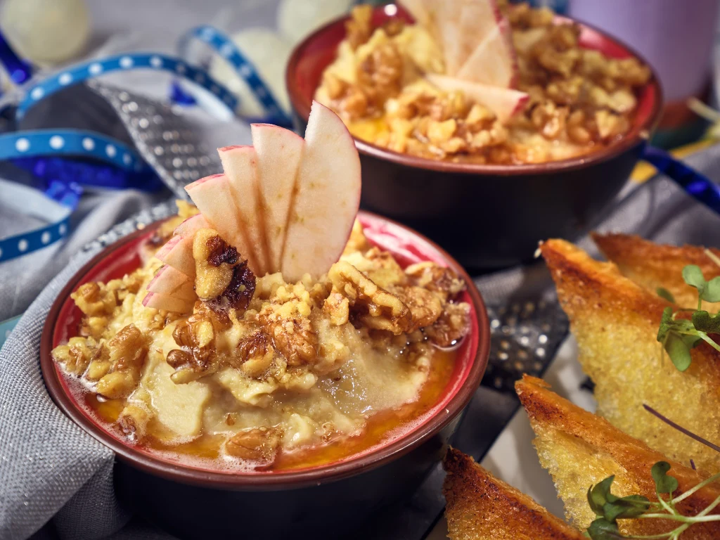 Miska kremowego deseru posypana orzechami włoskimi oraz plasterkami jabłka, obok chrupiące grzanki ozdobione zielonymi kiełkami, w tle kolorowe wstążki.