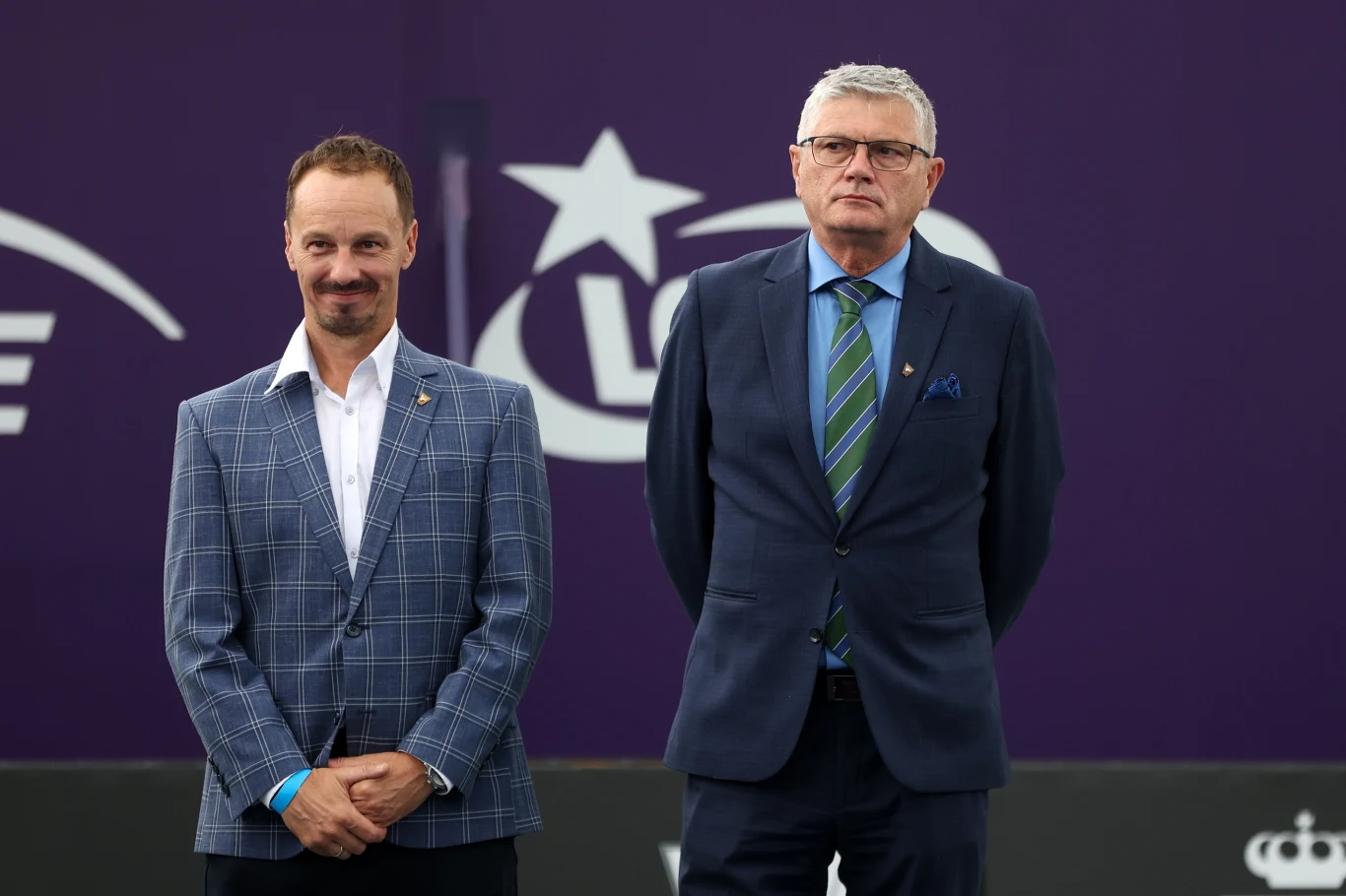 Prezes Dariusz Łukaszewski (z prawej), obok wiceprezes Radosław Szymanik Dwóch mężczyzn w garniturach stoi na tle fioletowej ścianki ozdobionej białymi znakami graficznymi, w tym gwiazdą i literami. Jeden z nich ma jasnobrązowe włosy i wąsy, drugi siwe włosy i okulary.