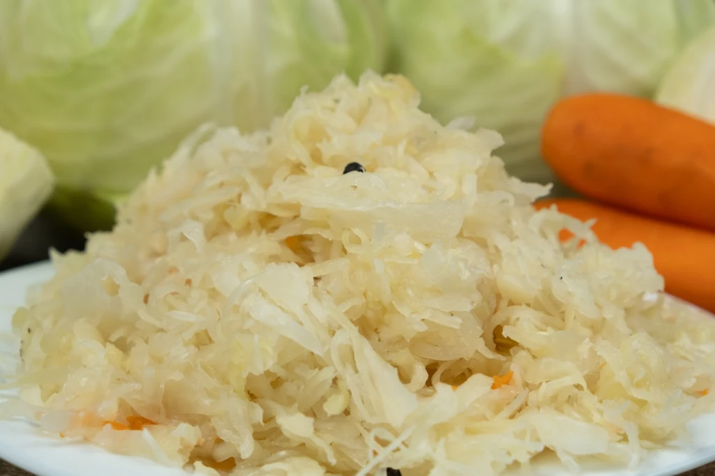 Kapusta kiszona to bogactwo witaminy C i naturalnych probiotyków. Jej wpływ na zdrowie jest nieoceniony Porcja kiszonej kapusty ułożona na białym talerzu, z widocznymi kawałkami marchwi oraz ziarnami czarnego pieprzu, w tle główki świeżej kapusty i całe marchewki.