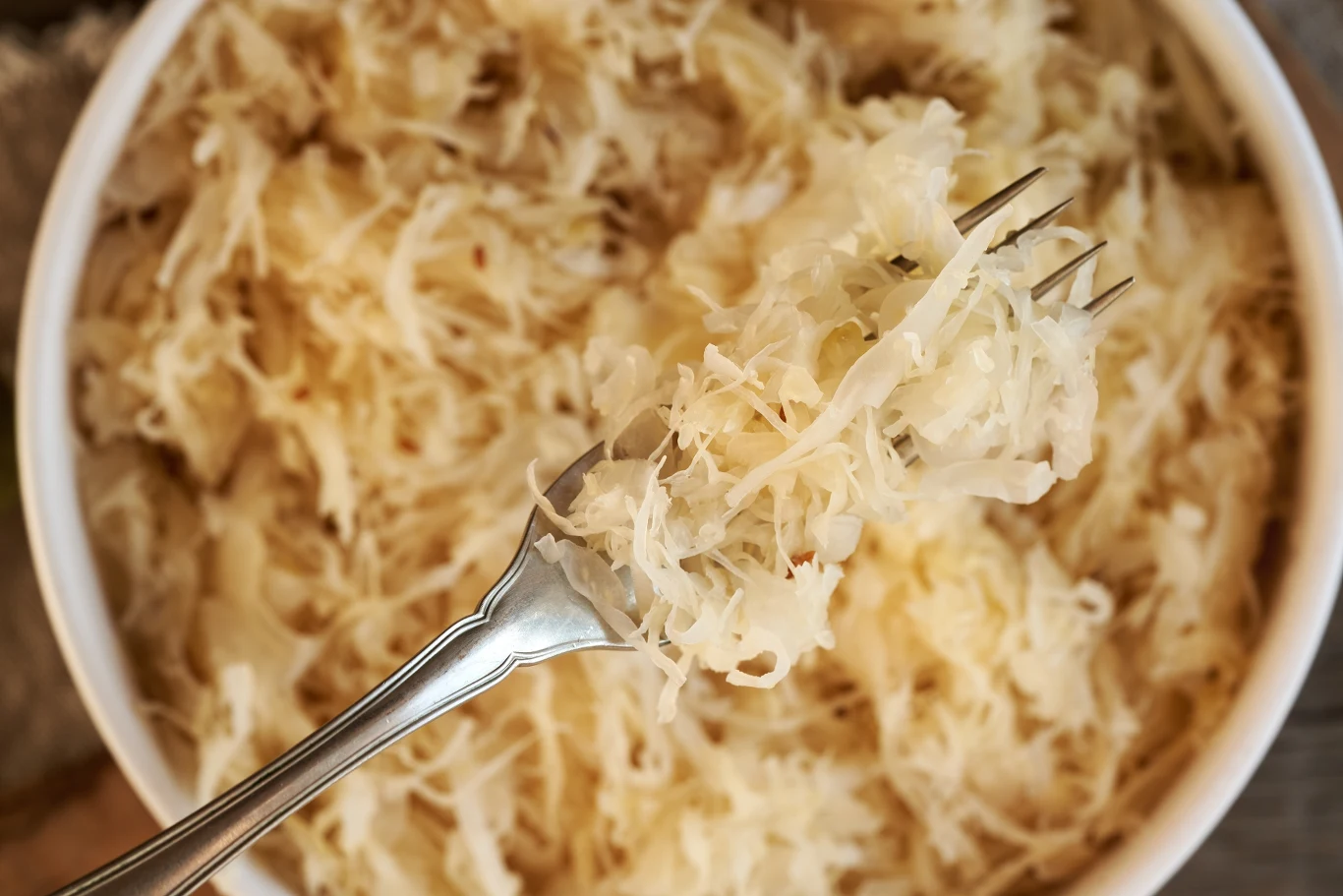 Kiszonki to samo zdrowie. W sezonie jesienno-zimowym powinny być obowiązkowe w diecie Szklana miska wypełniona kiszoną kapustą z widelcem nabierającym porcję kapusty, charakterystyczna struktura i kolor fermentowanego warzywa są dobrze widoczne.