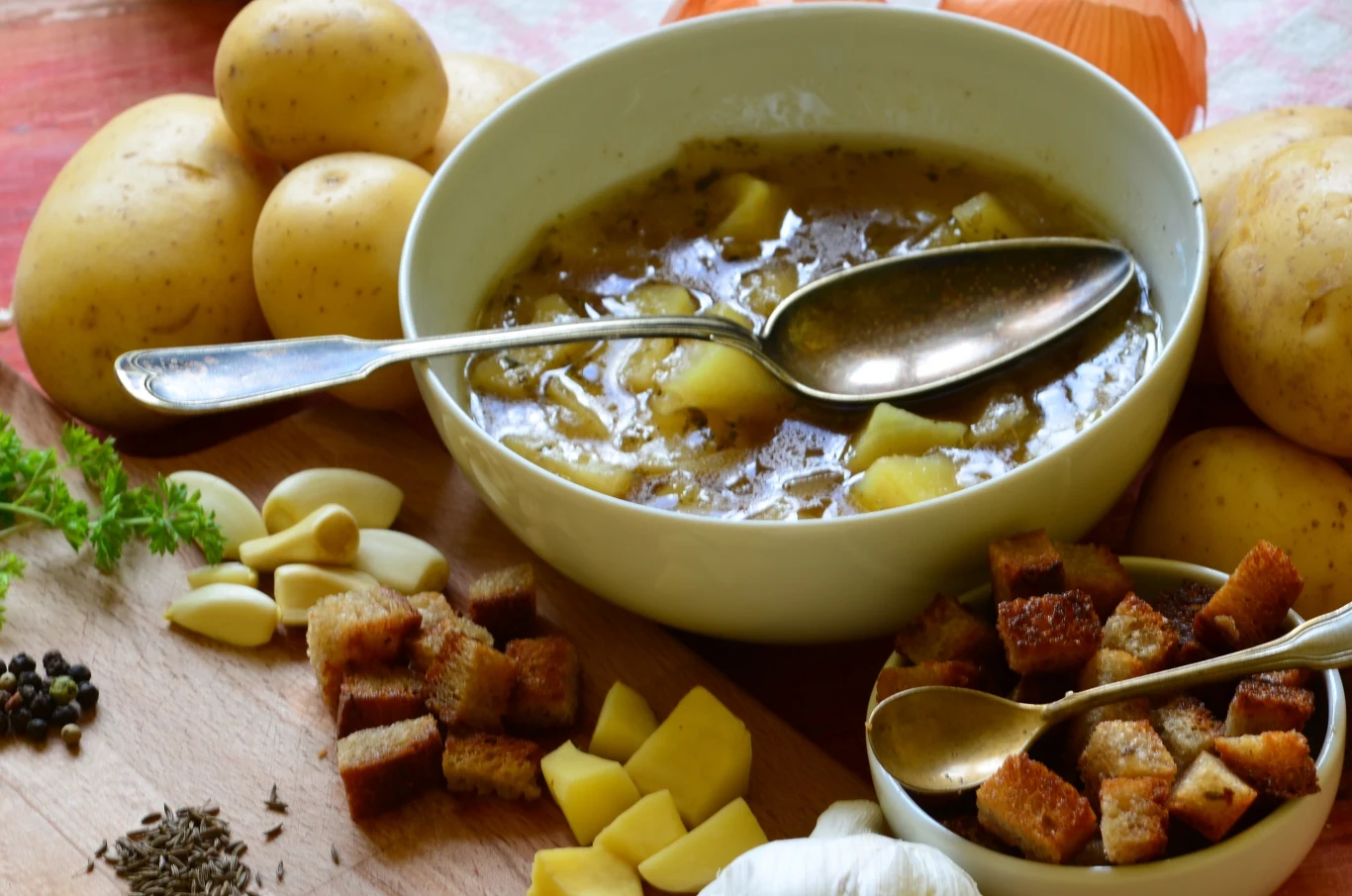 Miseczka aromatycznej zupy ziemniaczanej z widocznymi kawałkami ziemniaków, obok niej na desce kuchennej znajdują się czosnek, pieprz, grzanki oraz cebula i zioła. W tle widoczne całe ziemniaki.