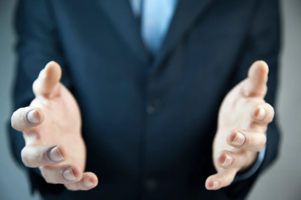 Wyciągnięte do przodu dłonie osoby ubrane w garnitur, rozluźnione palce, delikatny nieostry kadr skupiający się na gestykulacji dłońmi, rozmyte tło.