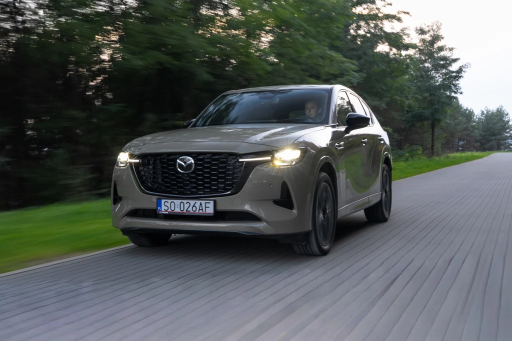 Mazda CX-60 jest obecna na rynku już od 3 lat Jadący po asfaltowej, leśnej drodze samochód osobowy marki Mazda z włączonymi światłami, na tle zieleni i sylwetek drzew, widoczny z przodu pod lekkim kątem.