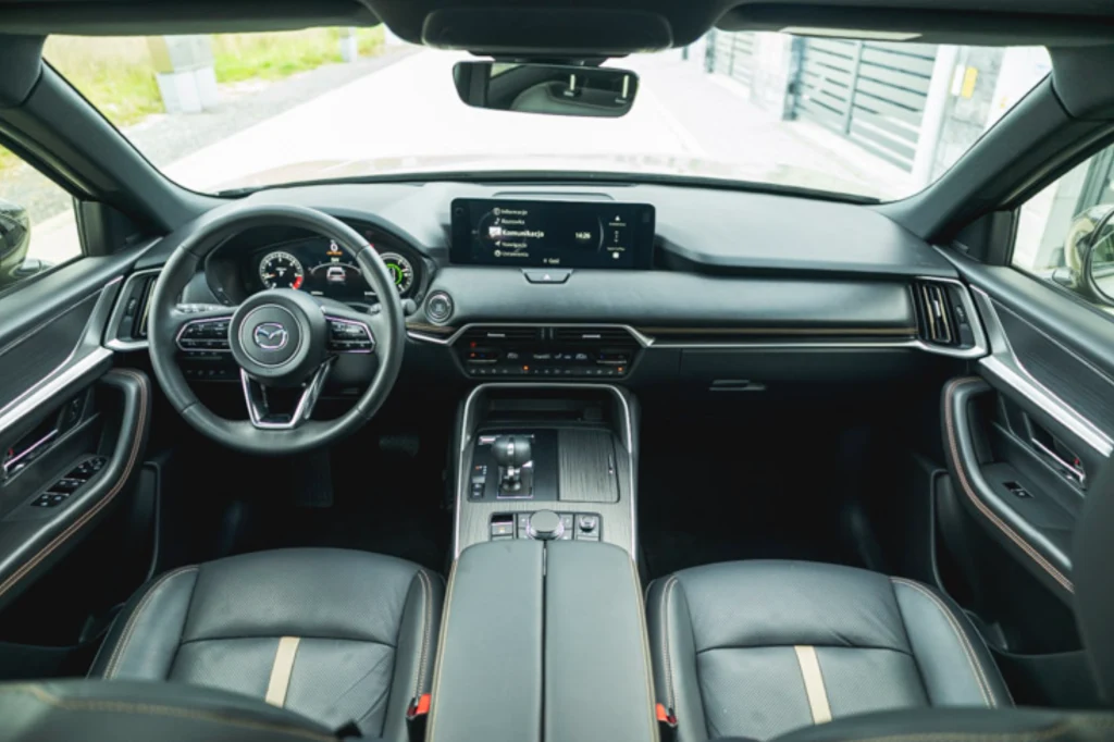 Mazda CX-60 ma dość minimalistyczne wnętrze, ale nie zabrakło w nim fizycznych przycisków Nowoczesne wnętrze samochodu marki Mazda z dużym ekranem systemu multimedialnego na desce rozdzielczej, skórzanymi siedzeniami i eleganckim wykończeniem kokpitu.
