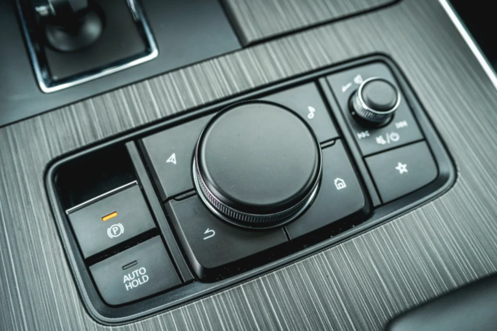 Centralne pokrętło z przyciskami pozwalającymi szybko wybrać najważniejsze opcje kiedyś było popularnym rozwiązaniem, obecnie to duży wyróżnik CX-60 Panel sterowania systemem multimedialnym samochodu z dużym pokrętłem centralnym, kilkoma przyciskami funkcyjnymi oraz teksturą imitującą szczotkowane aluminium.