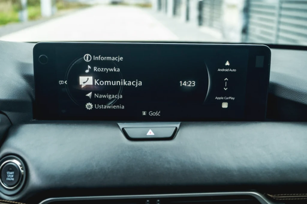 System multimedialny w Mazdzie CX-60 ma bardzo minimalistyczną grafikę i jest prosty w obsłudze, choć nie wszystko rozplanowano w nim intuicyjnie Panel multimedialny wewnątrz samochodu z wyświetlaczem ukazującym menu ustawień, na którym widoczne są opcje takie jak komunikacja, rozrywka, nawigacja i ustawienia. Na ekranie widoczna jest godzina oraz przyciski funkcyjne Android Auto i Apple CarPlay.