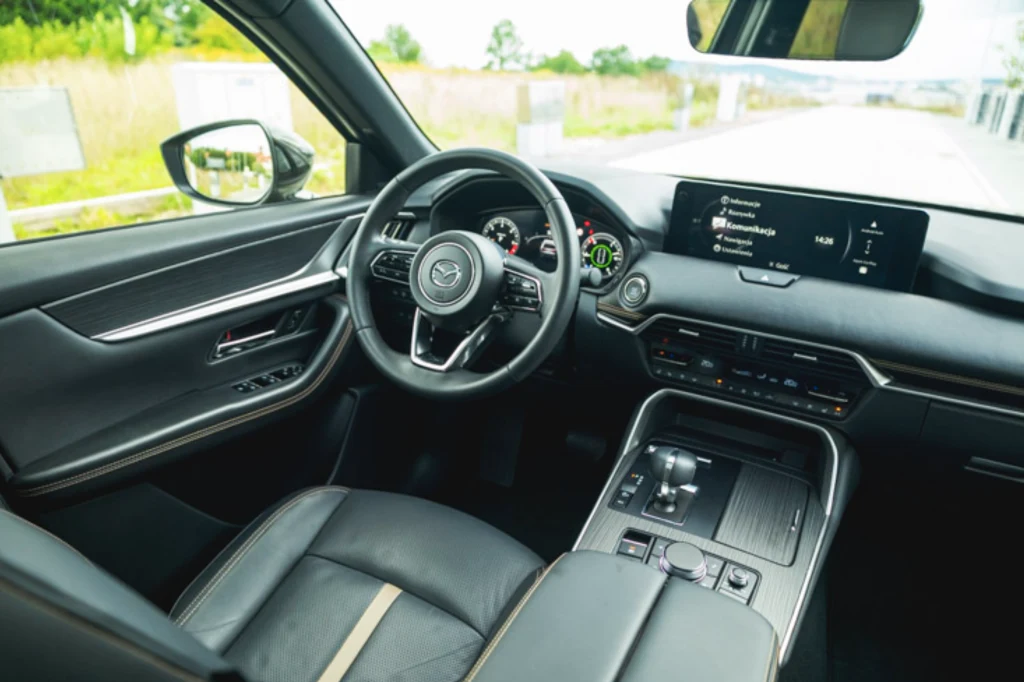 Skóra Nappa z kontrastowymi przeszyciami oraz winylowe i metalizowane wstawki bardzo poprawiają odbiór wnętrza CX-60 Nowoczesne wnętrze samochodu z kierownicą marki Mazda, elektronicznym panelem zegarów, dużym ekranem multimedialnym i luksusowymi skórzanymi fotelami. Widok na otoczenie przez boczne okno samochodu.