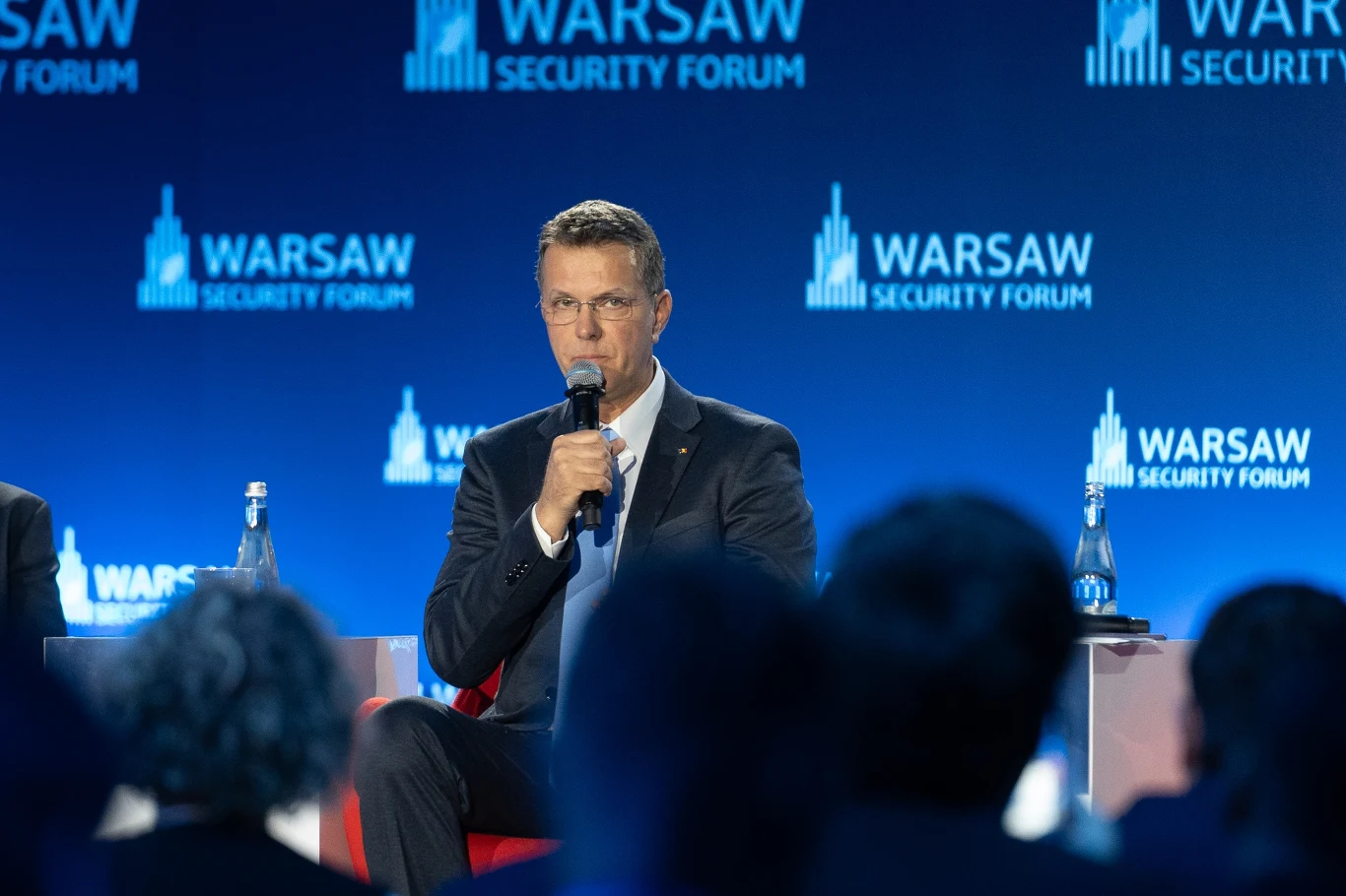 Minister obrony Ionut Mosteanu podał się do dymisji Mężczyzna w garniturze siedzi na scenie, trzyma mikrofon i mówi podczas konferencji Warsaw Security Forum, na tle niebieskiej scenografii z logo wydarzenia.