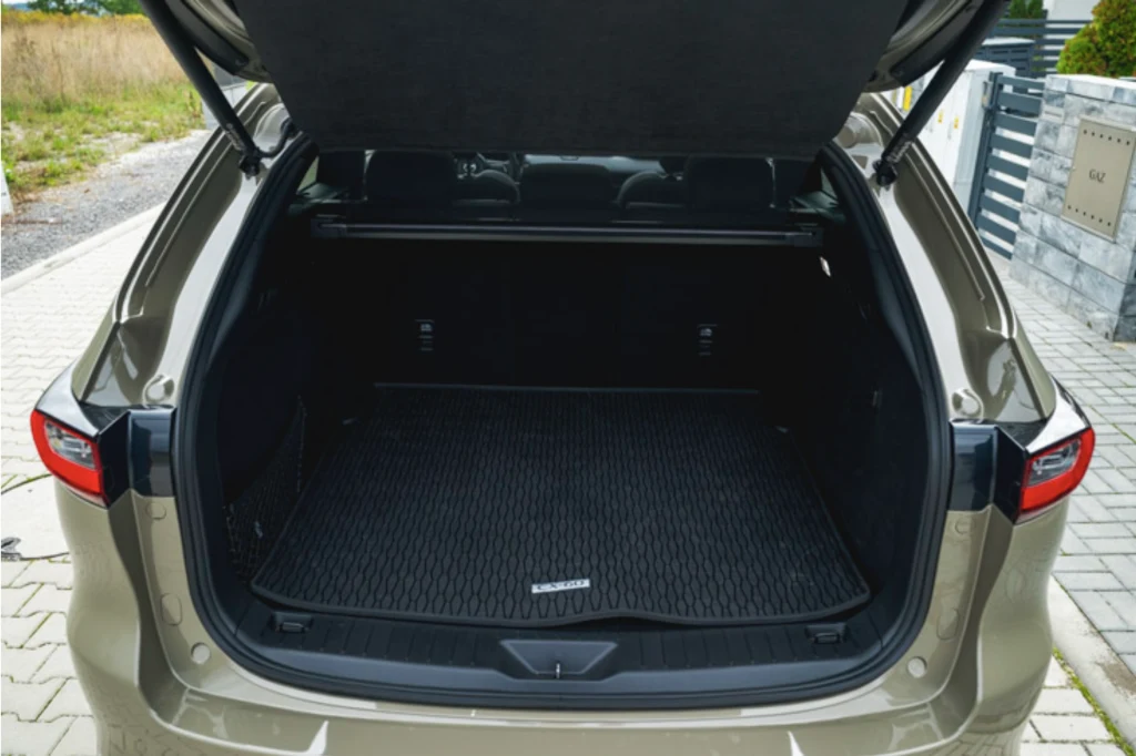 Bagażnik CX-60 ma pojemność 570 l Otwarty bagażnik samochodu osobowego zaparkowanego na brukowanej nawierzchni, we wnętrzu widoczna czysta, gumowa mata ochronna, wokół samochodu fragment ogrodzenia i zieleni.