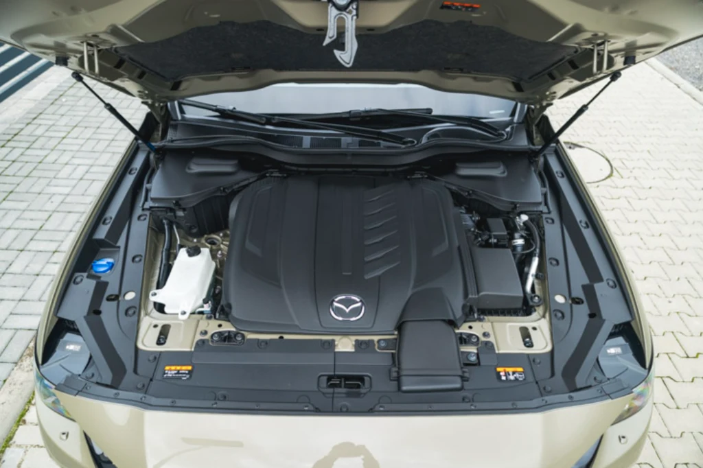 Mazda CX-60 napędzana jest sześciocylindrowym, rzędowym dieslem o pojemności 3,3 l Otwarta maska samochodu osobowego z wyeksponowanym silnikiem marki Mazda oraz innymi elementami komory silnika, widoczne detale techniczne i estetyczne obudowy.