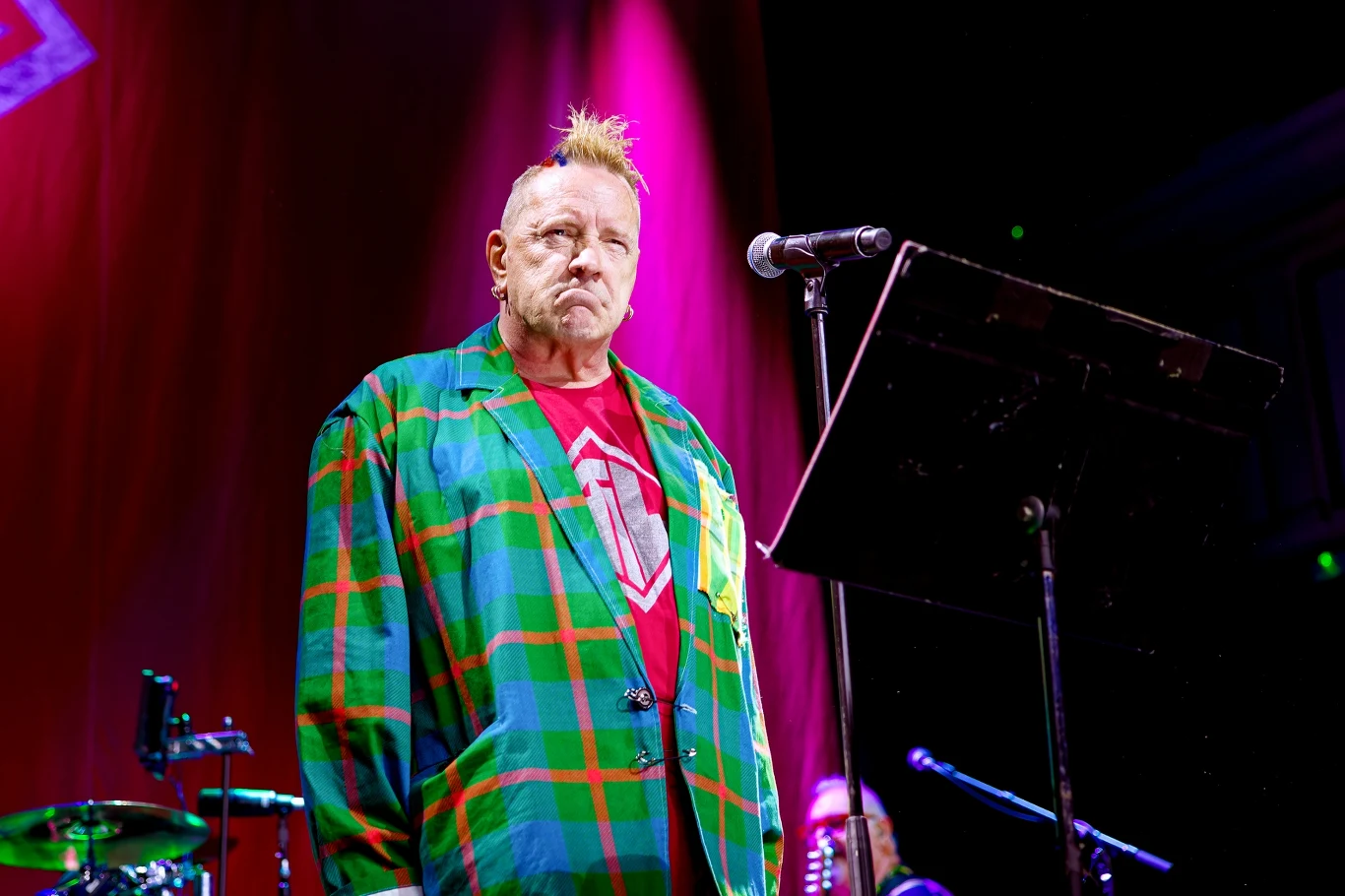 John Lydon stoi na czele grupy PiL Starszy mężczyzna w kolorowej marynarce i czerwonej koszulce stoi przed mikrofonem na scenie, w tle oświetlenie na różowo i elementy perkusji. Mężczyzna ma charakterystyczną fryzurę z irokezem oraz poważny wyraz twarzy.