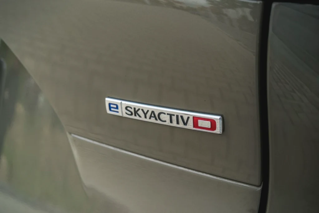 Diesel Mazdy, pomimo sporej pojemności, okazuje się oszczędny. Wspomaga go układ miękkiej hybrydy Metalowy emblemat samochodu z napisem „e SKYACTIV D” umieszczony na błotniku auta o szarobrązowym kolorze lakieru.