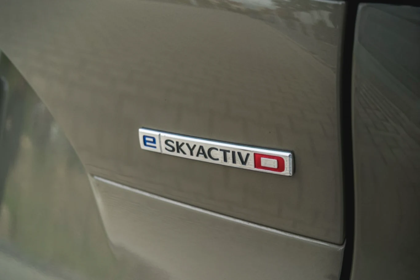 Diesel Mazdy, pomimo sporej pojemności, okazuje się oszczędny. Wspomaga go układ miękkiej hybrydy Metalowy emblemat samochodu z napisem „e SKYACTIV D” umieszczony na błotniku auta o szarobrązowym kolorze lakieru.