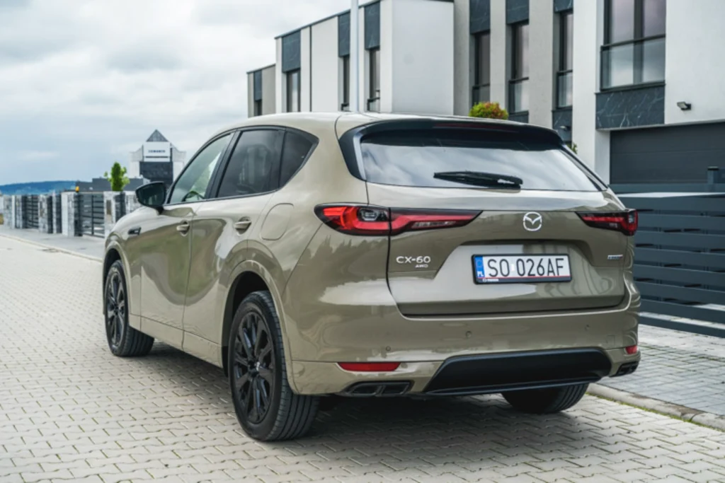 Testowana CX-60 pokryta była ciekawym lakierem o nazwie "cyrkonowy piasek", który dołączył do oferty w ubiegłym roku Nowoczesny samochód SUV marki Mazda CX-60 w beżowym kolorze, zaparkowany na brukowanej drodze przed szeregiem nowoczesnych budynków mieszkalnych.