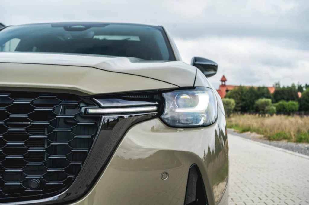Mazda CX-60 Zbliżenie na przednią część samochodu osobowego z włączonym reflektorem LED, wyrazisty grill, elementy maski i fragment szyby, w tle rozmyte drzewa oraz budynek.