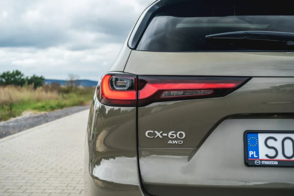 Mazda CX-60 Tylna część samochodu Mazda CX-60 AWD w kolorze beżowym na tle brukowanej drogi i krajobrazu z niewyraźną roślinnością, widoczne tylne światło oraz polska tablica rejestracyjna.