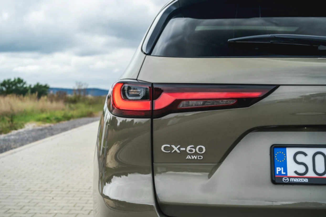 Mazda CX-60 Tylna część samochodu Mazda CX-60 AWD w kolorze beżowym na tle brukowanej drogi i krajobrazu z niewyraźną roślinnością, widoczne tylne światło oraz polska tablica rejestracyjna.
