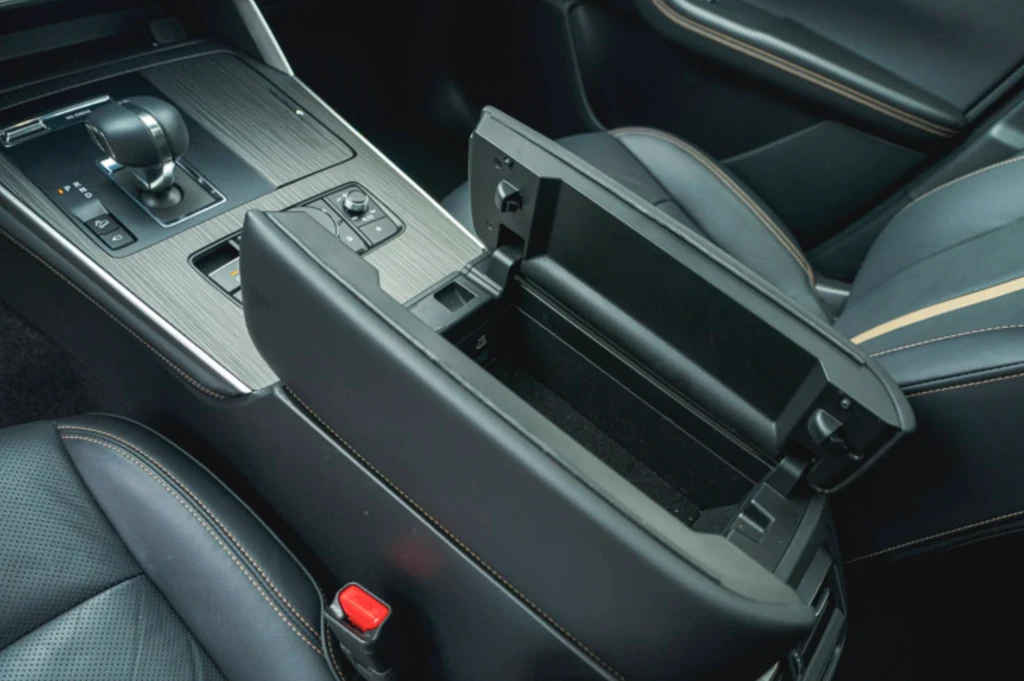 Mazda CX-60 Środek nowoczesnego samochodu z otwartym schowkiem podłokietnika, widoczną dźwignią zmiany biegów, panelem sterowania i czarnymi skórzanymi fotelami z przeszyciami.