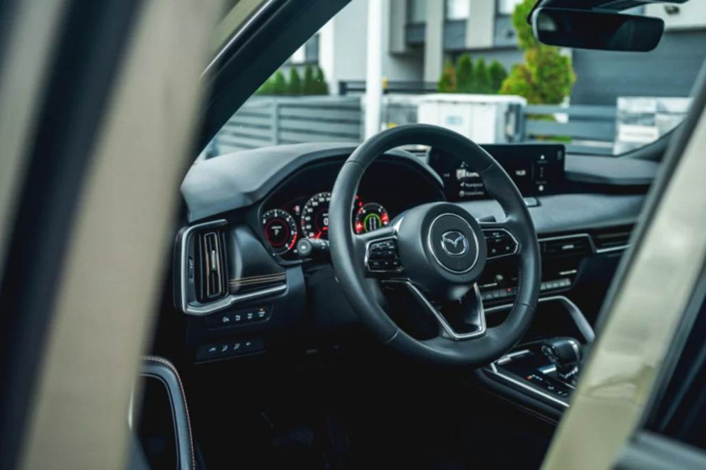Mazda CX-60 Kokpit nowoczesnego samochodu Mazda z widoczną kierownicą, deską rozdzielczą i cyfrowymi wskaźnikami, po lewej stronie panel sterowania światłami, po prawej ekran systemu multimedialnego, w tle elementy miejskiej zabudowy.