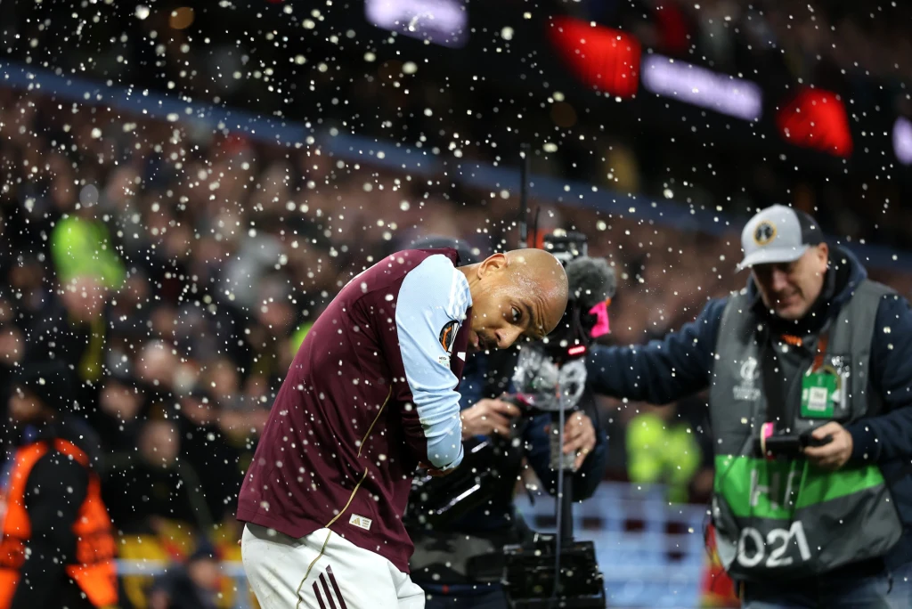 Piłkarz w bordowej koszulce uderzony przez padający deszcz lub napój podczas meczu, w tle tłum na trybunach oraz operator z kamerą i pracownik obsługi w kurtce odblaskowej.