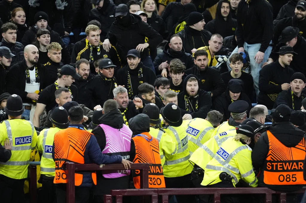 Grupa kibiców ubranych głównie na czarno i żółto w czasie konfliktu lub zamieszania na trybunach, oddzielona od policji i ochroniarzy ustawionych w kamizelkach odblaskowych.