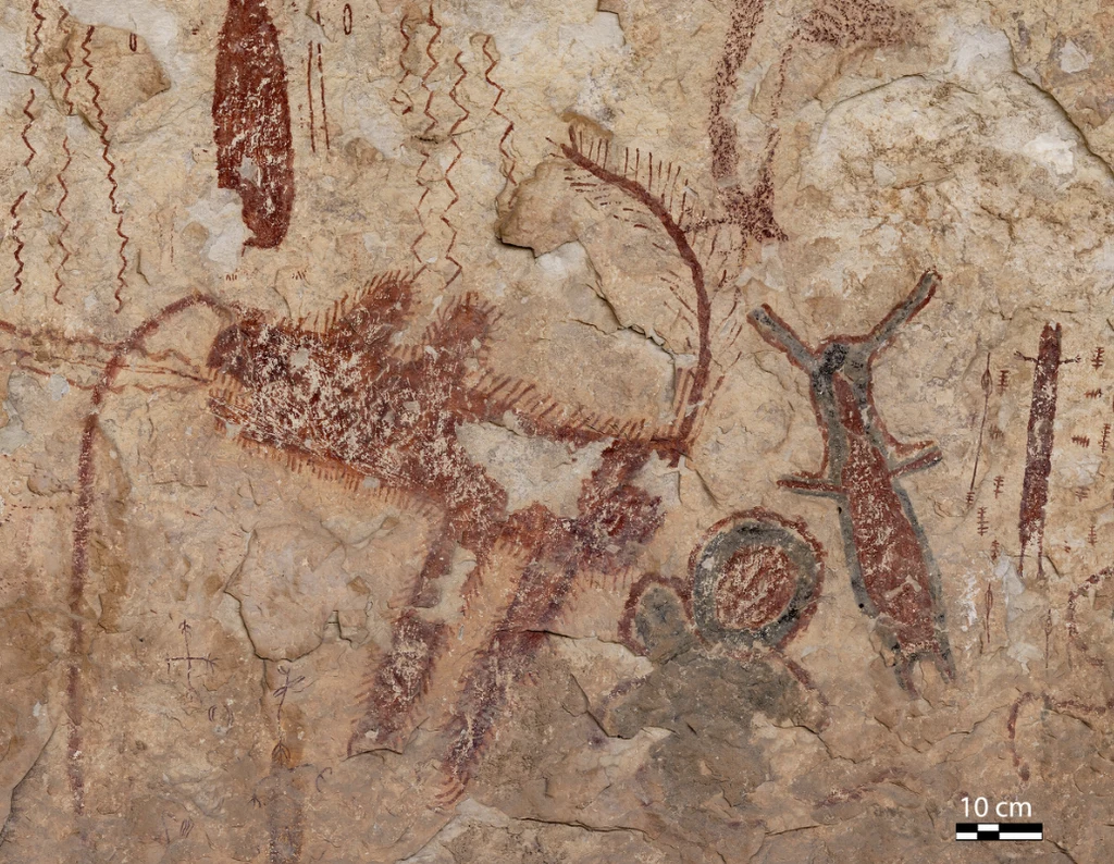 Motywy malowideł naskalnych wykonane czerwoną i czarną farbą na kamiennej powierzchni, przedstawiające figury ludzi, zwierząt oraz abstrakcyjne linie faliste i kształty geometryczne.