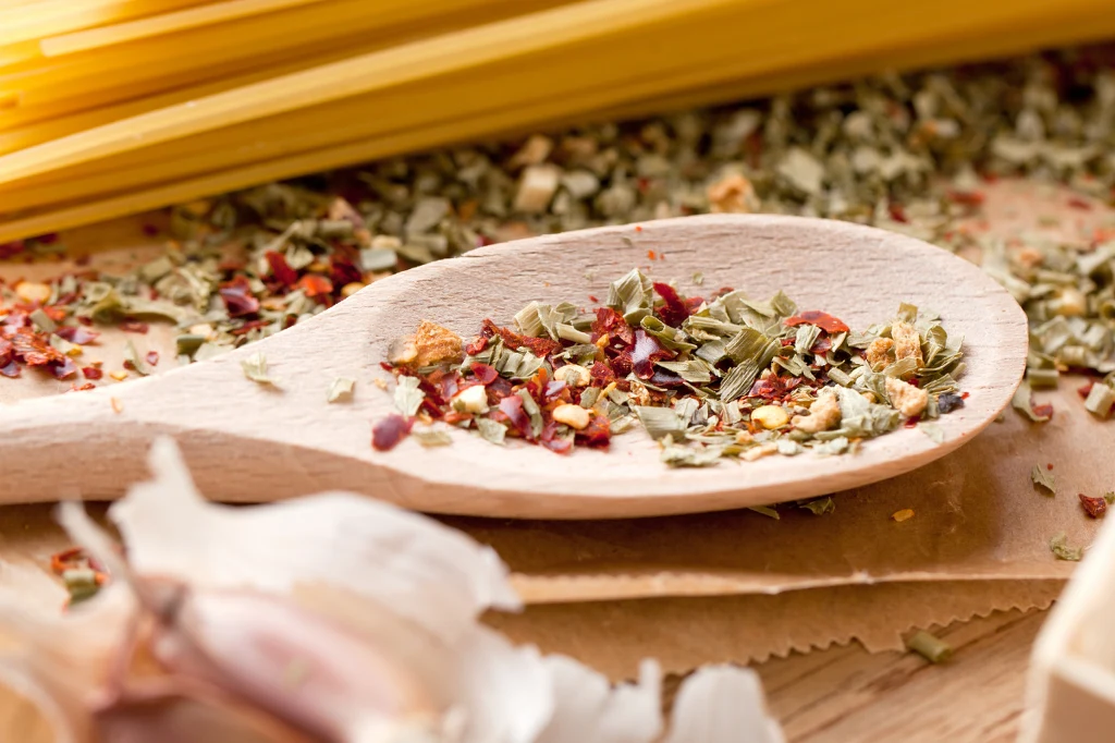 Włoska przyprawa z oregano, peperoncino i czosnkiem to patent na zdrowie i szczupłą sylwetkę Drewniana łyżka wypełniona mieszanką suszonych ziół i przypraw, w tle żółty makaron oraz porozrzucane składniki na drewnianej powierzchni.