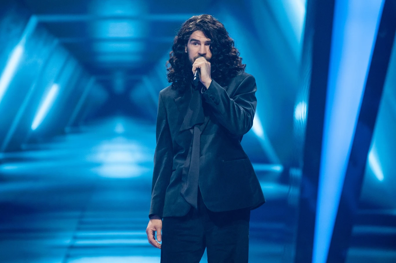 Dominik Czuż odpadł w półfinale "The Voice of Poland" Mężczyzna z długimi, kręconymi włosami ubrany w ciemny garnitur śpiewa do mikrofonu na futurystycznie oświetlonej scenie z niebieskimi światłami i geometrycznym tłem.