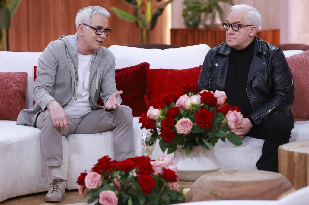 Robert Janowski doprowadził Wojciecha Bardowskiego do wygranej w "The Voice Senior" Dwóch starszych mężczyzn siedzi na białych sofie i rozmawia w jasnym, przytulnym wnętrzu z dekoracjami kwiatowymi z czerwonych i różowych róż oraz zieleni w tle. Jeden z mężczyzn ubrany jest w szary dres i okulary, drugi w czarną skórzaną kurtkę i okul...
