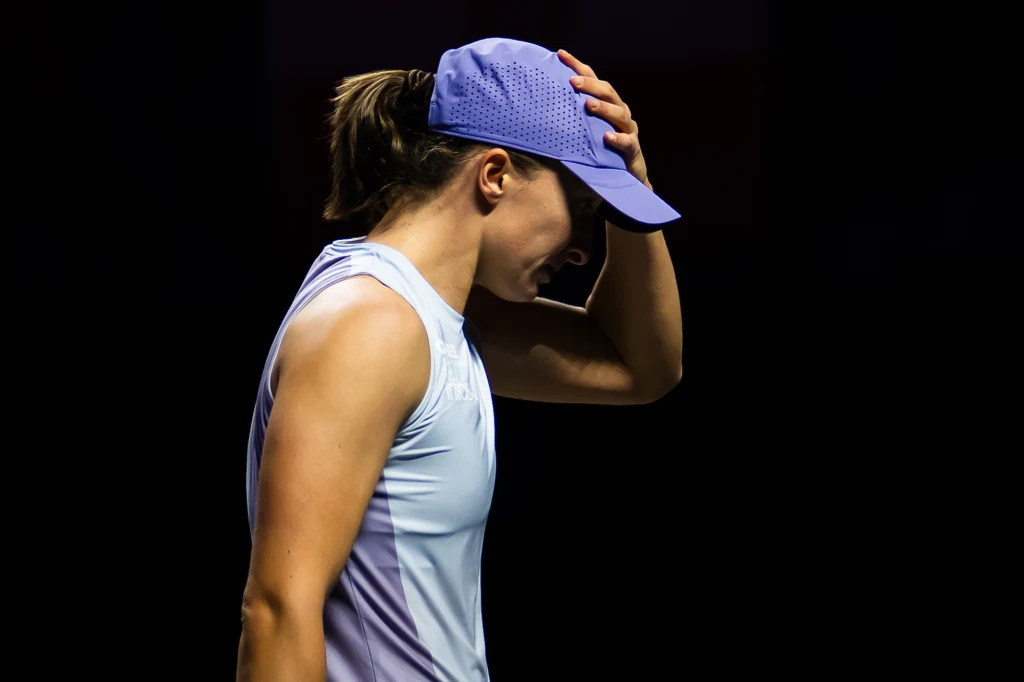 Iga Świątek - wiceliderka rankingu WTA na korcie Kobieta ubrana w sportowy top i czapkę z daszkiem, trzymająca dłoń na czole w geście zmęczenia lub stresu, na ciemnym tle.