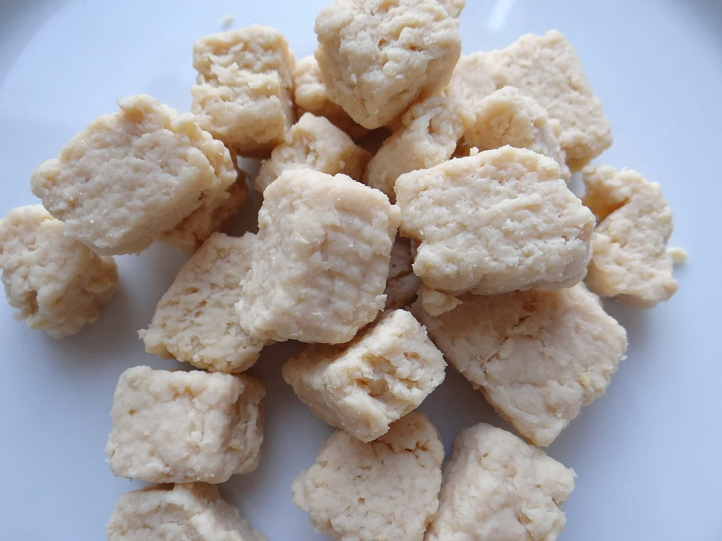 Kostki jasnobeżowej tekstury ułożone na białym tle, przypominające suchy produkt spożywczy, prawdopodobnie kawałki sojowe lub tofu.