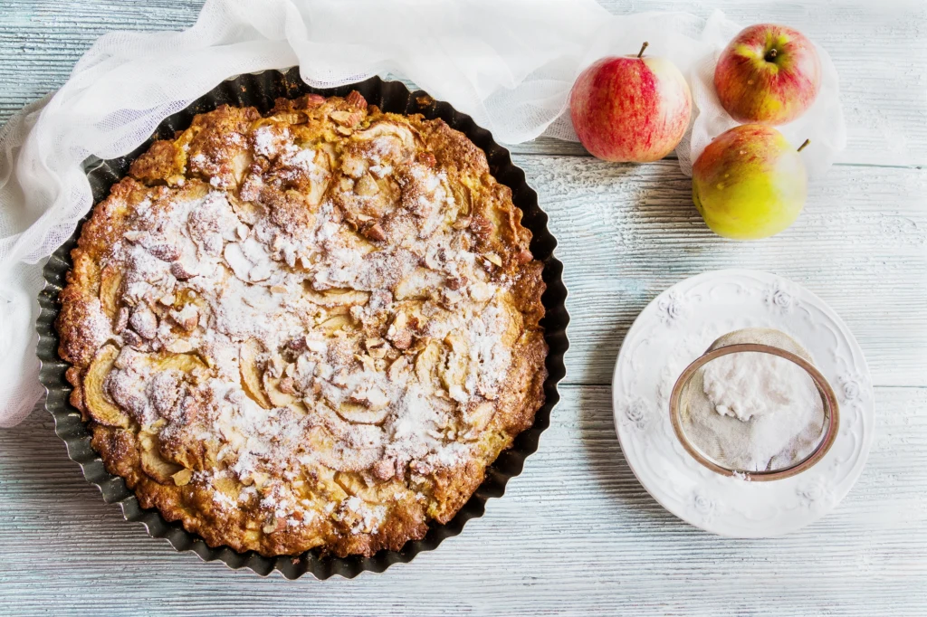 Szarlotka posypana cukrem pudrem w okrągłej formie do pieczenia na białym, drewnianym stole z trzema czerwono-żółtymi jabłkami i talerzykiem z cukrem pudrem obok.