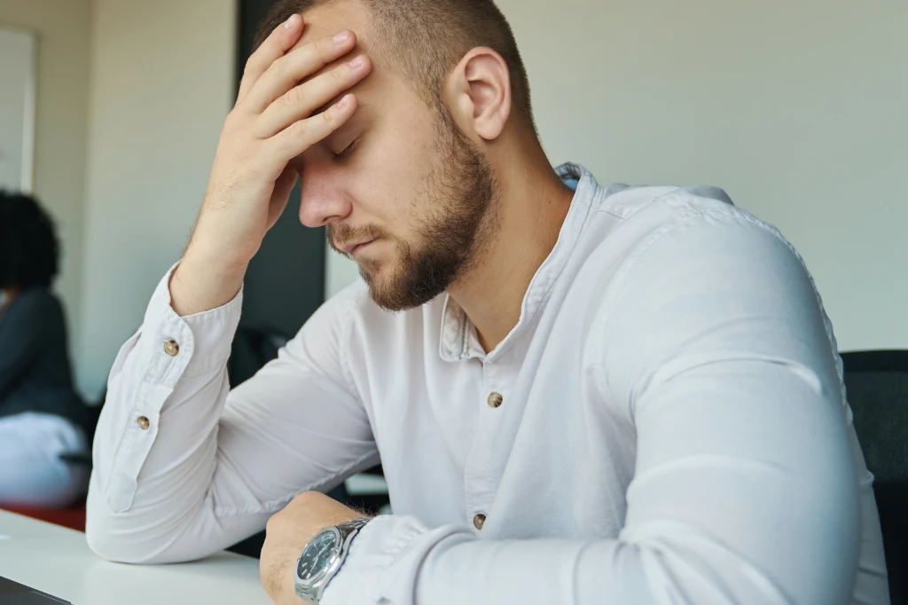 Objawy glejaka mogą przypominać padaczkę lub udar Mężczyzna z brodą w jasnej koszuli opiera głowę o dłoń z zamkniętymi oczami, sprawiając wrażenie zmęczenia, stresu lub zmartwienia, siedząc przy stole w biurze.