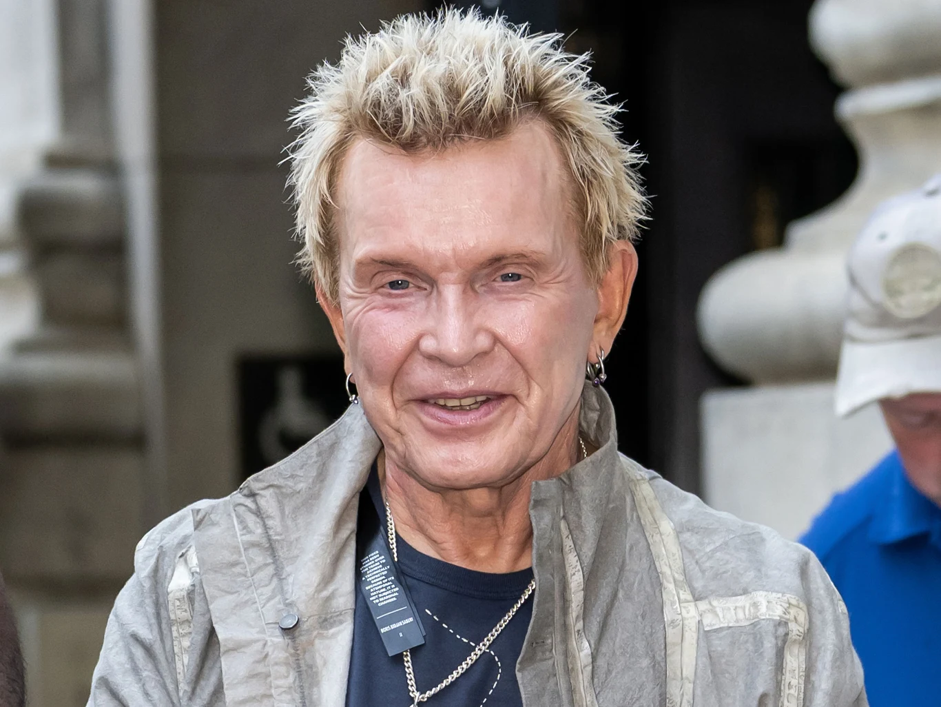Billy Idol 30 listopada kończy 70 lat Uśmiechnięty mężczyzna o jasnych, krótkich włosach z zadbaną fryzurą, ubrany w szarą kurtkę i ciemny podkoszulek, na szyi ma kilka srebrnych łańcuszków. W tle fragment miejskiej architektury i niewyraźne postacie.