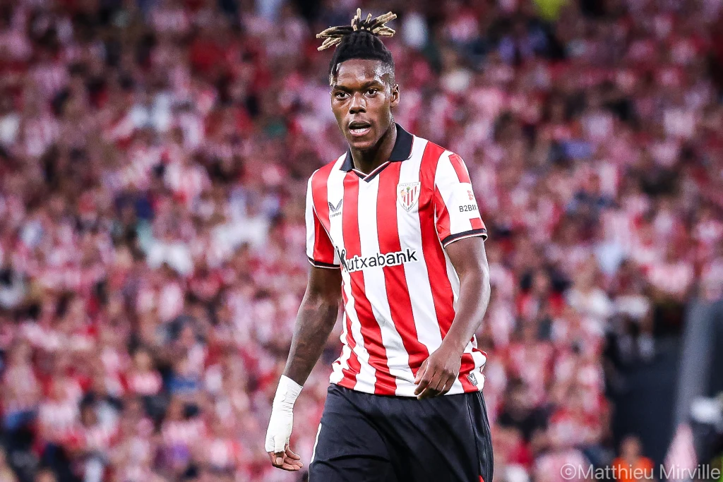 Piłkarz w czerwono-białej koszulce Athletic Bilbao na stadionie, koncentrujący się podczas gry, w tle rozmyta publiczność w trybunach.