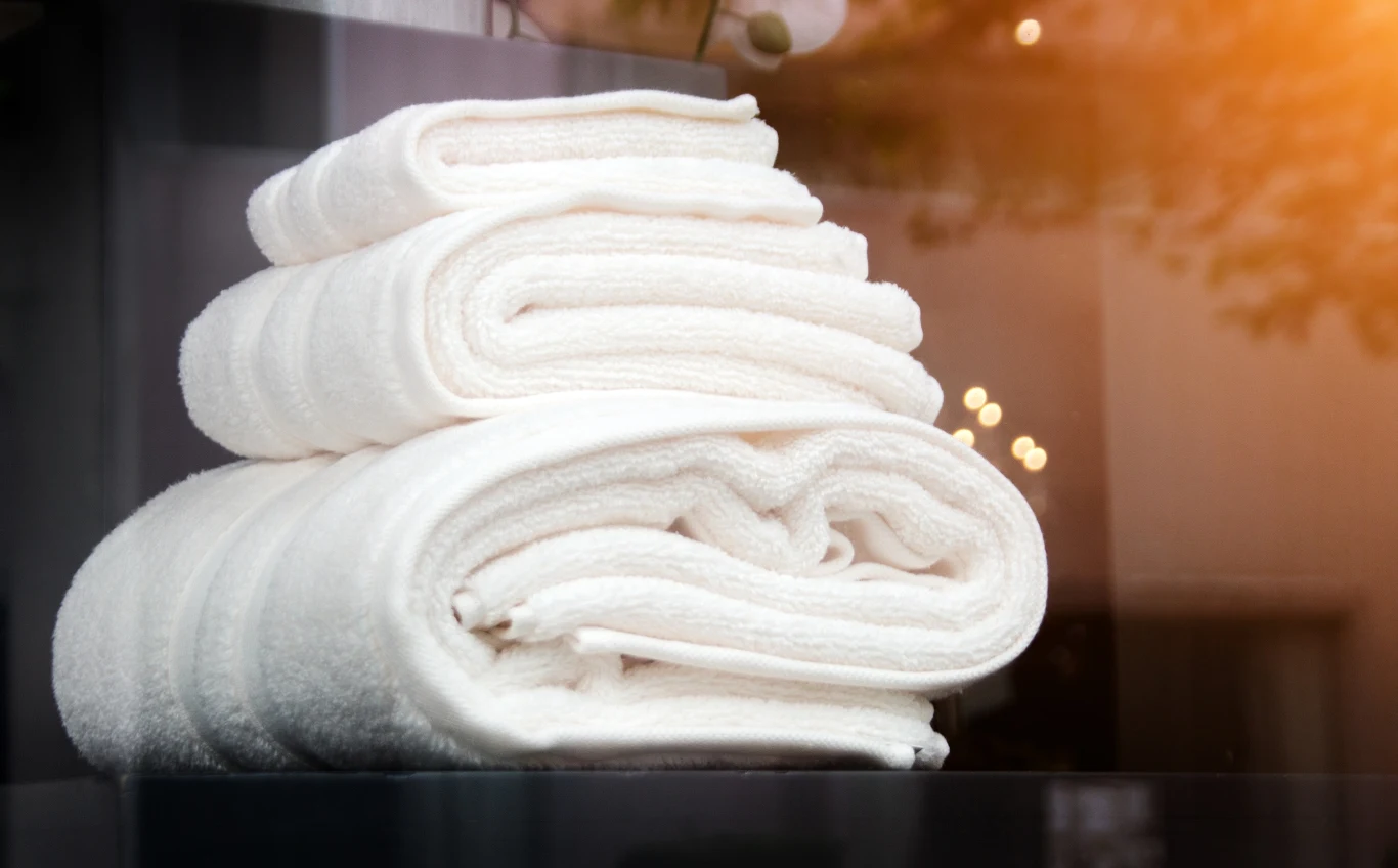 Ręczniki będą miękkie i puszyste Stos jasnobiałych, schludnie złożonych ręczników leżących na szklanej powierzchni, w tle widać delikatne refleksy światła i rozmyte elementy imitujące spokojną atmosferę.