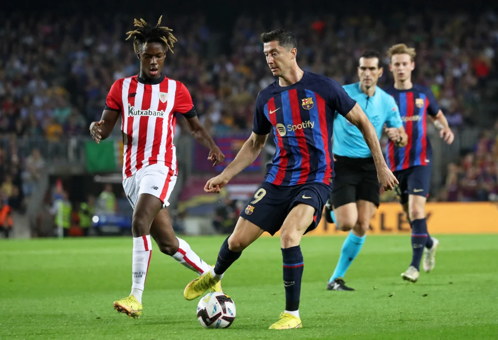 Piłkarz w koszulce FC Barcelony kontroluje piłkę na murawie podczas meczu, zbliżając się do rywala w barwach Athletic Bilbao. W tle widoczni są inni zawodnicy, sędzia oraz kibice na trybunach.