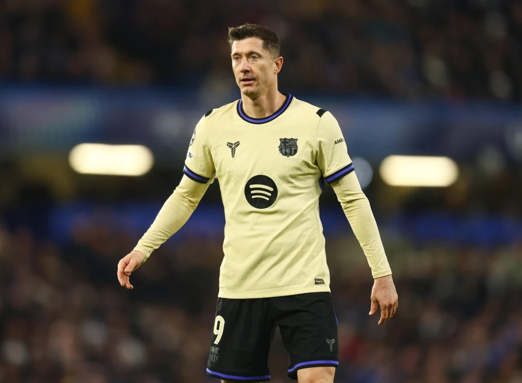 Piłkarz w jasnej koszulce z numerem 9 na boisku, w tle rozmazana publiczność stadionowa, logo drużyny oraz sponsora widoczne na koszulce.