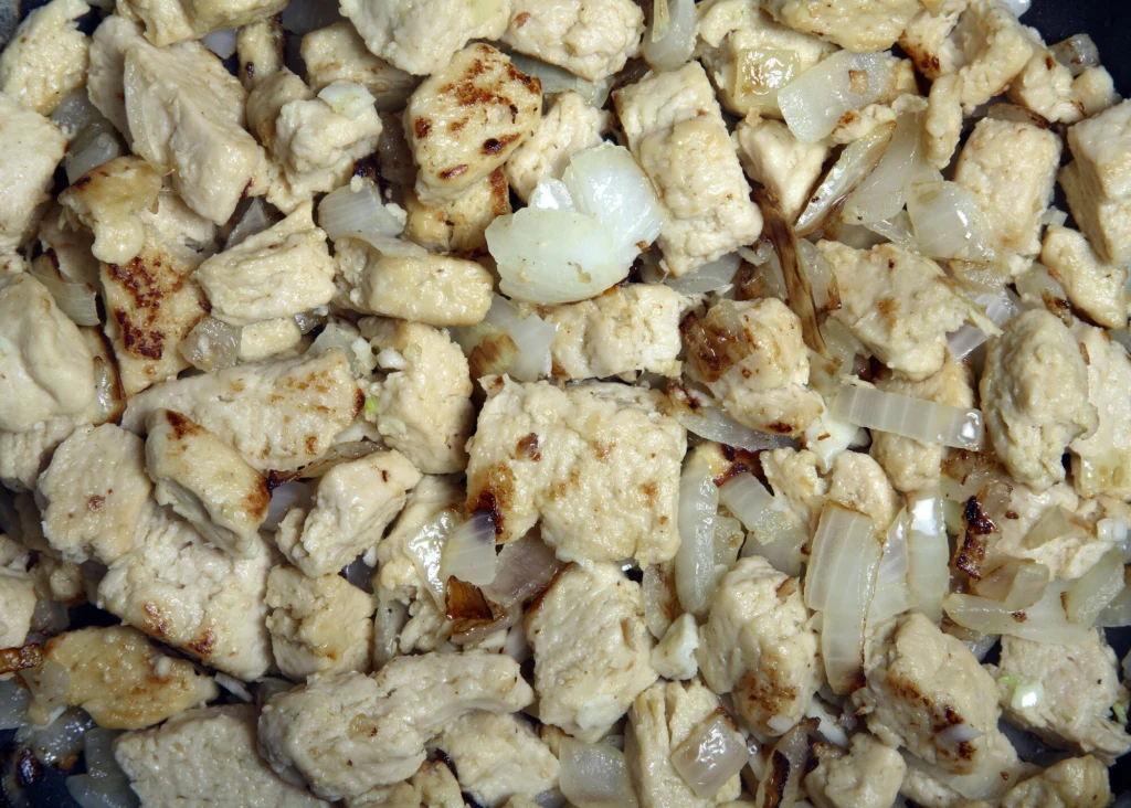 Smażone kawałki mięsa drobiowego z wyraźnie widoczną, zeszkloną cebulą, podane na patelni, przyrumienione na złoty kolor.