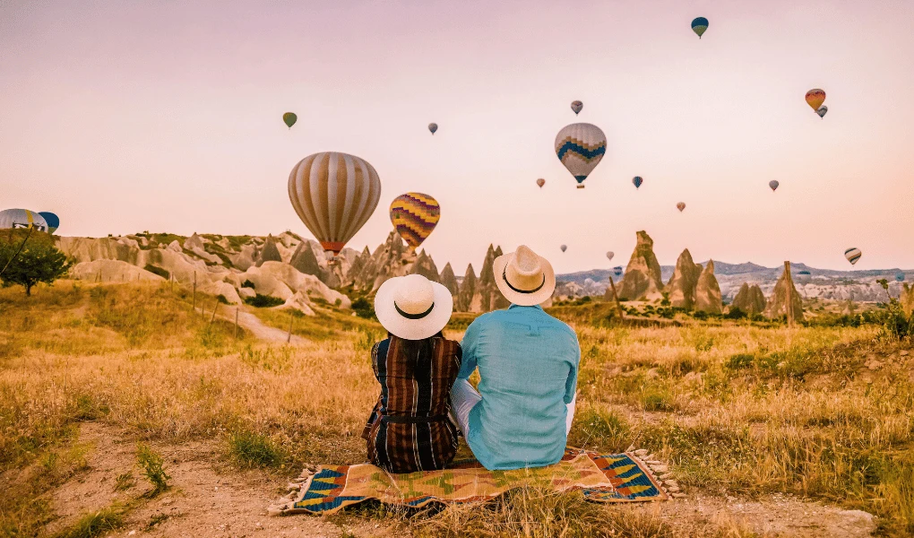 Co robić w Turcji poza hotelem? Dwie osoby w kapeluszach siedzące na kocu na suchym polu, zwrócone tyłem do obiektywu, podziwiające unoszące się balony na tle skalistych formacji i różowego nieba.