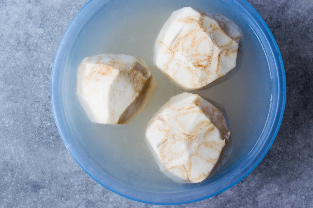 Zamiast gotować, spróbujcie upiec seler. Jest nie tylko pyszny, ale i zdrowy Trzy obrane bulwy selera zanurzone w wodzie w plastikowej misce na szarej powierzchni.