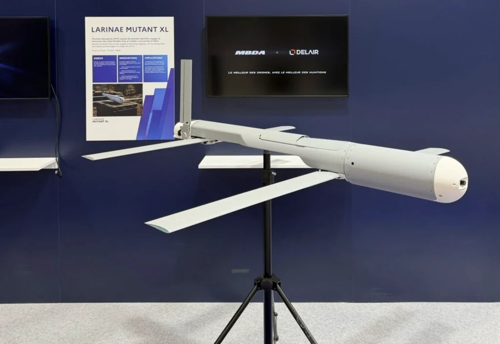 Bezzałogowy statek powietrzny z rozłożonymi skrzydłami ustawiony na statywie, w tle tablica informacyjna z nazwą Larinae Mutant XL oraz dwa ekrany z logotypami firm.