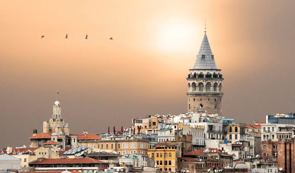 Zabytki w Turcji Stare zabytkowe budynki i charakterystyczna Wieża Galata o stożkowym dachu dominująca nad panoramą miasta, na tle ciepłego nieba z przelatującym stadem ptaków.