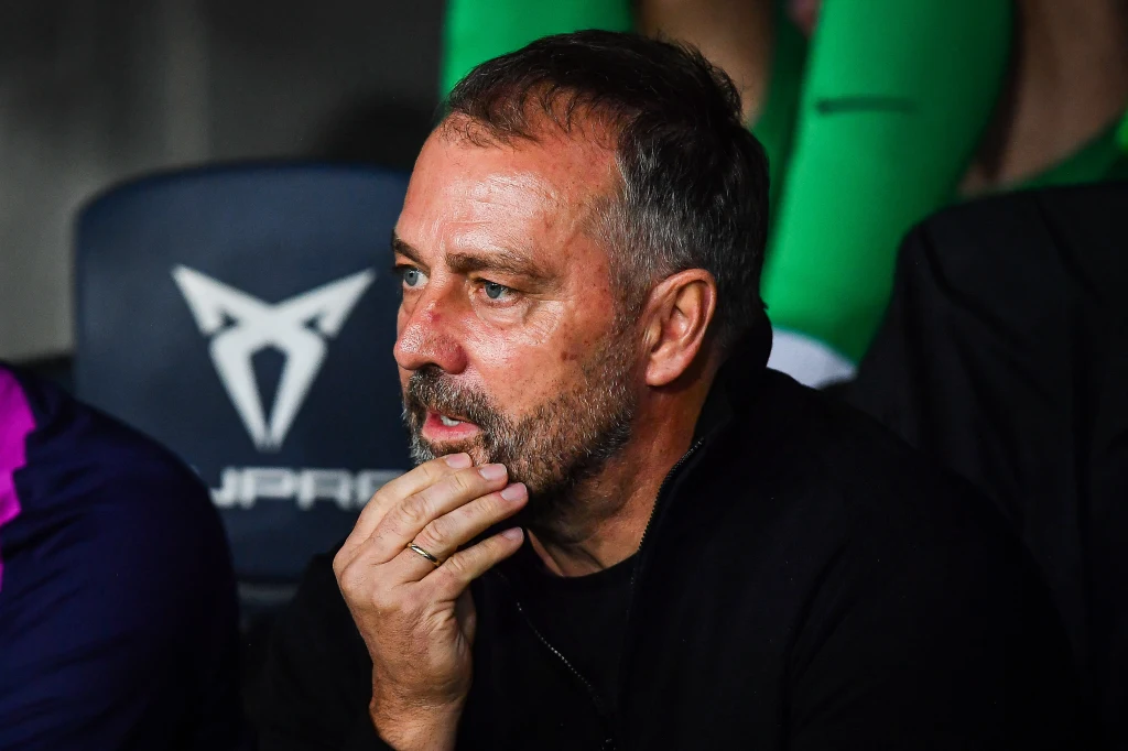 Mężczyzna z brodą siedzący na stadionowej ławce, opierający głowę o rękę w zamyśleniu, w tle ciemne krzesła i fragment zielonych spodni innej osoby.