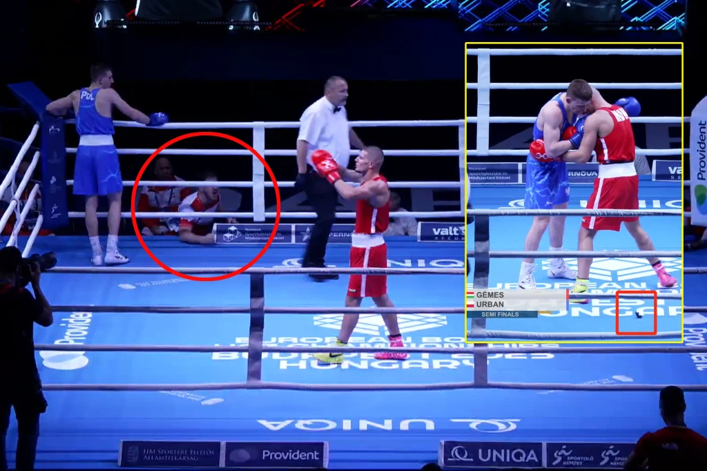 Dwóch bokserów na ringu podczas oficjalnej walki, jeden w niebieskim stroju siedzi przy linach, wyraźnie wyczerpany, drugi w czerwonym stoi zwycięsko uniesionymi rękami. W tle sędzia po środku ringu. Pojedyncze osoby widoczne poza ringiem.