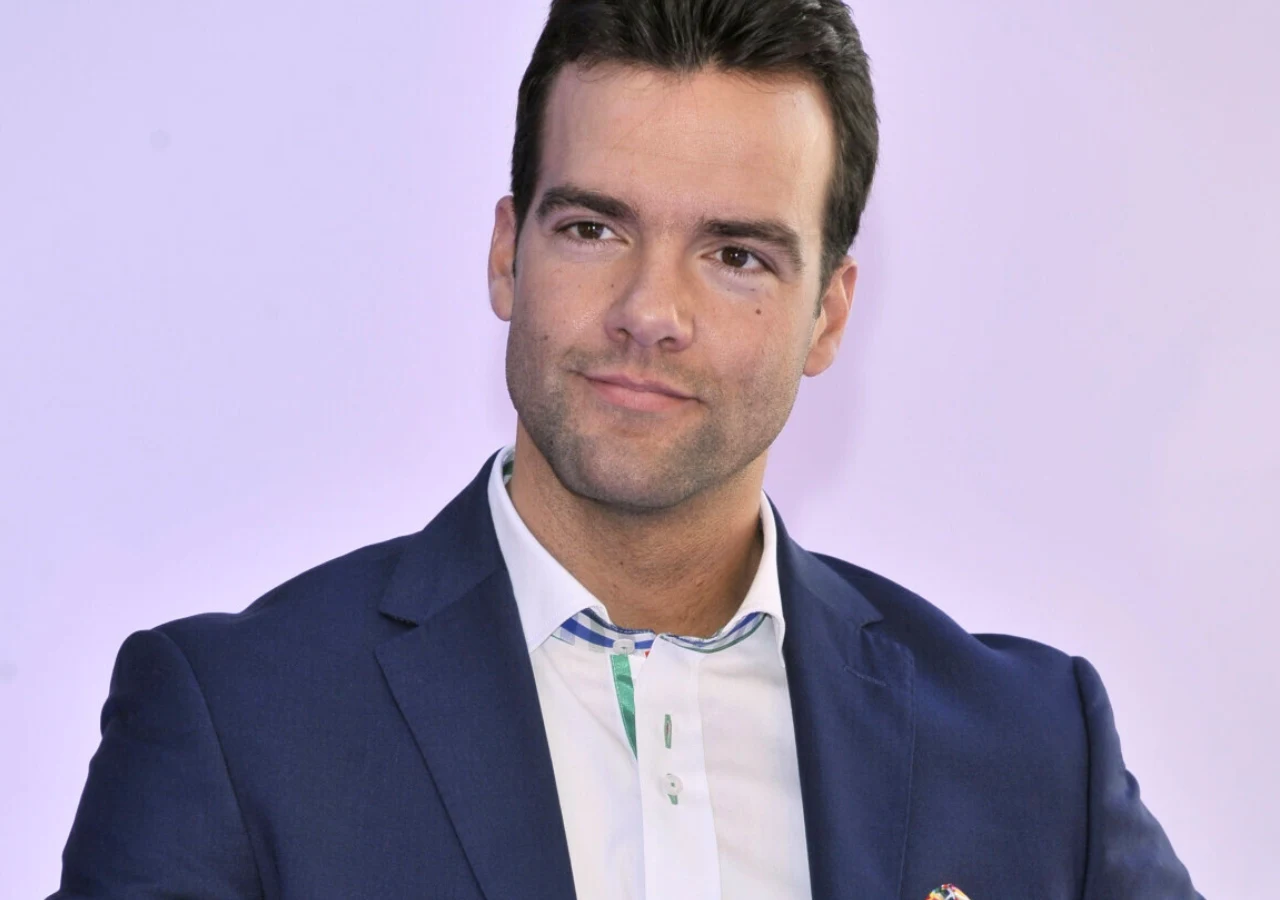 Conrado Moreno Mężczyzna z krótkimi, ciemnymi włosami, ubrany w granatowy garnitur i białą koszulę, spogląda lekko w bok z delikatnym uśmiechem, na jasnym tle.