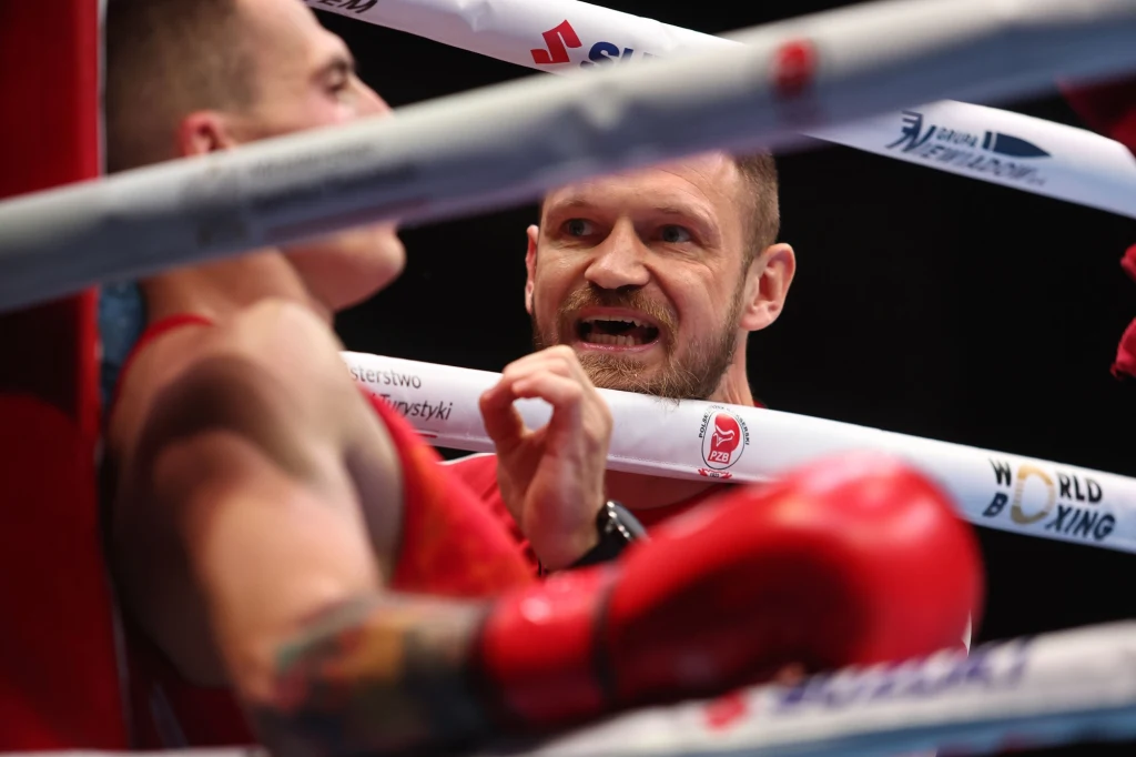 Trener udziela wskazówek bokserowi podczas przerwy w ringu; bokser siedzi na linach, uważnie słucha, obaj w sportowych strojach, w tle widoczne logo światowej organizacji bokserskiej.